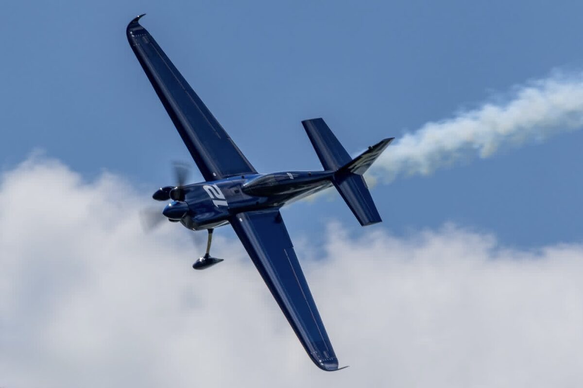 Topside view of the blue Zivko Edge 540 v3 racing number 12 banking hard with a smoke trail